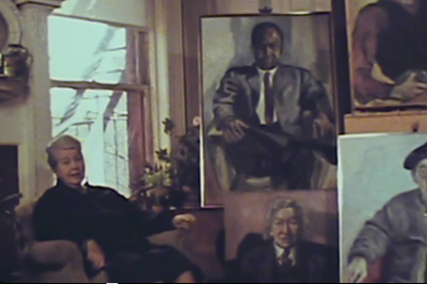 The artist Alice Neel sits on a chair surrounded by various portraits of people in pastel colors
